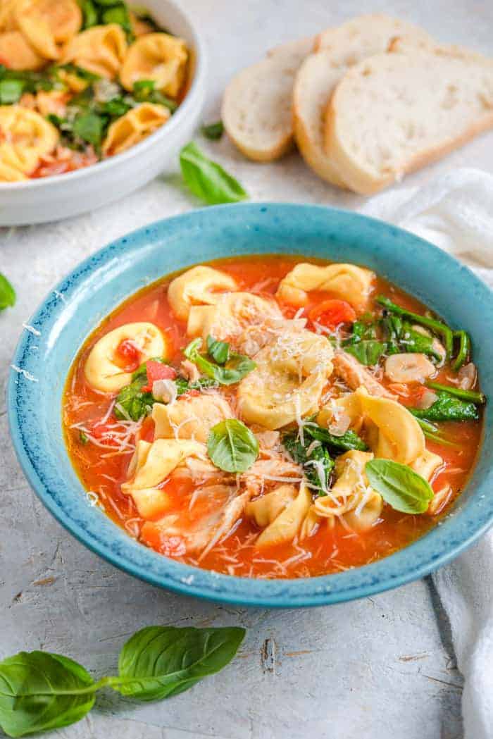 A side shot of tortellini soup in a blue bowl with bread in the background