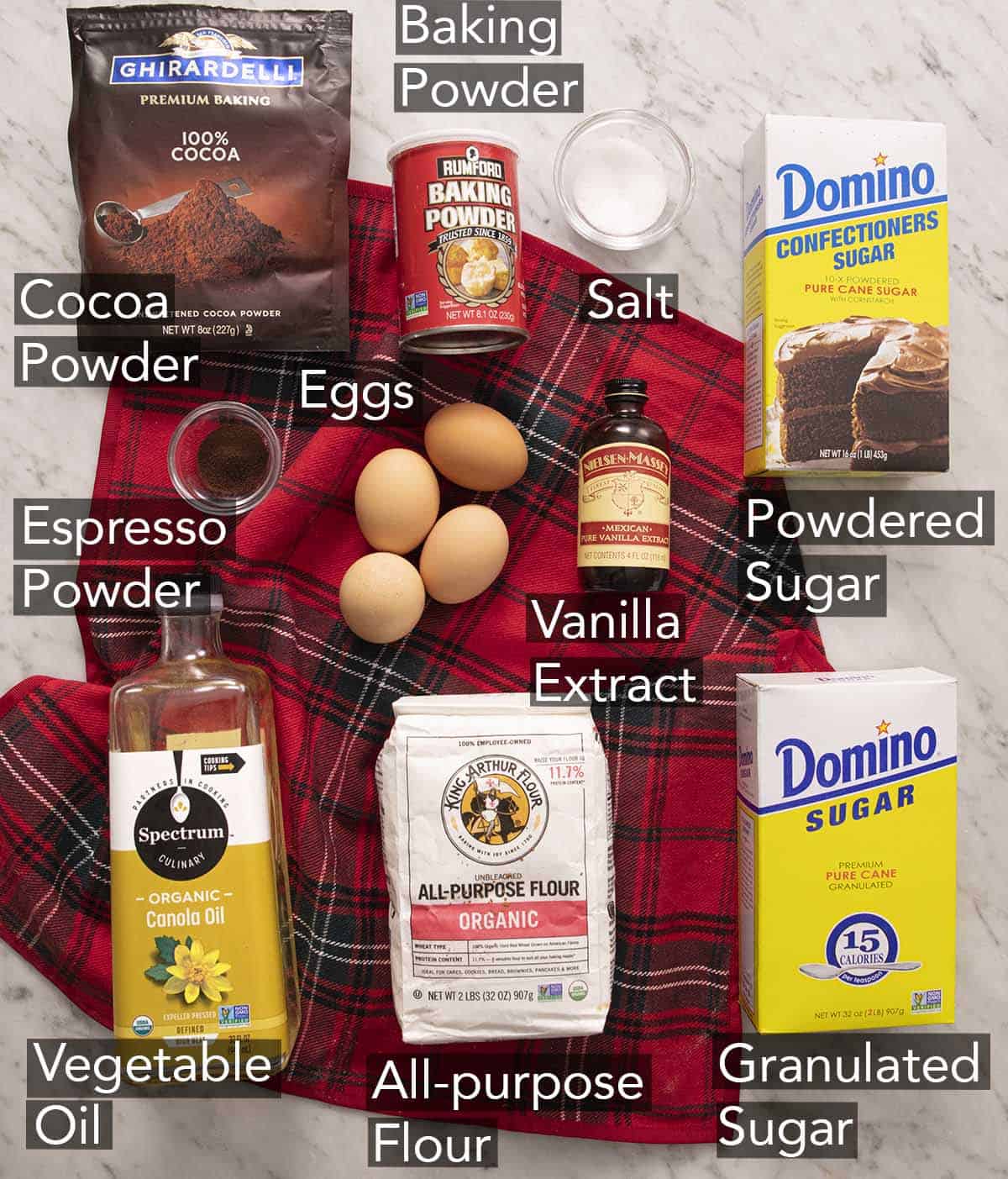 Ingredients to make chocolate crinkle cookies with text labels on a marble counter. 