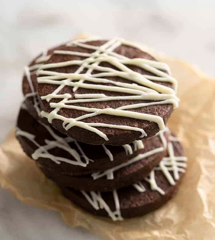 A stack of chocolate sugar cookies drizzled with chocolate on a piece of parchment paper. 