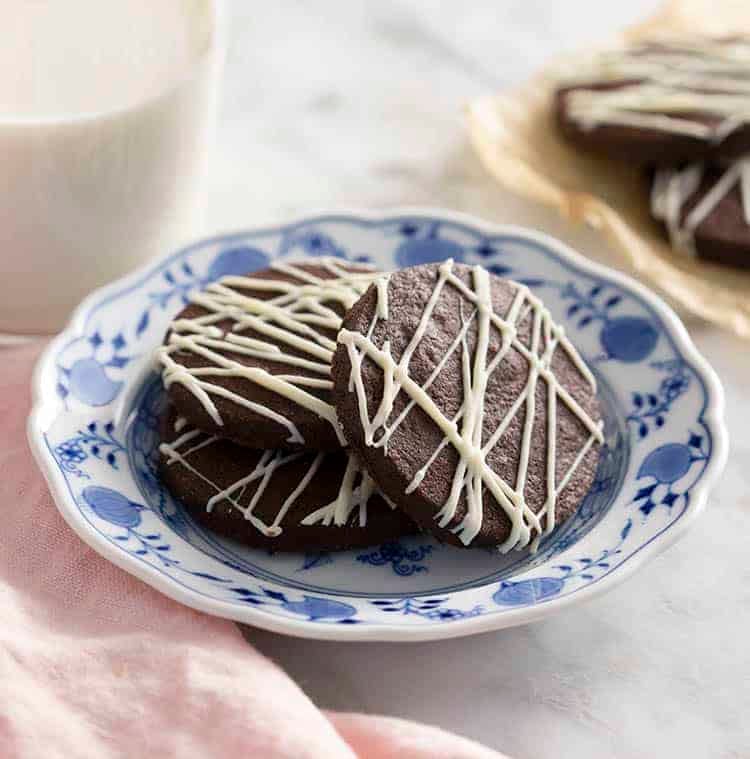 Chocolate sugar cookies drizzled with white chocolate on a blue and white plate next to a glass of milk.