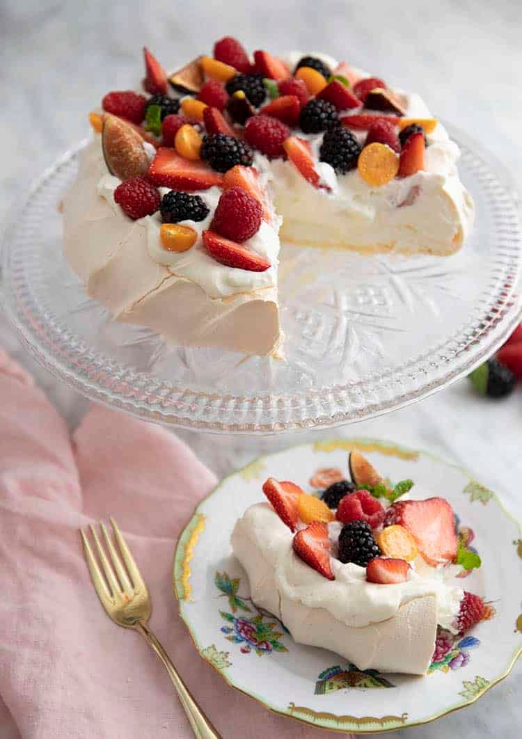 A pavlova on a crystal cake stand with a piece cut out in the foreground.