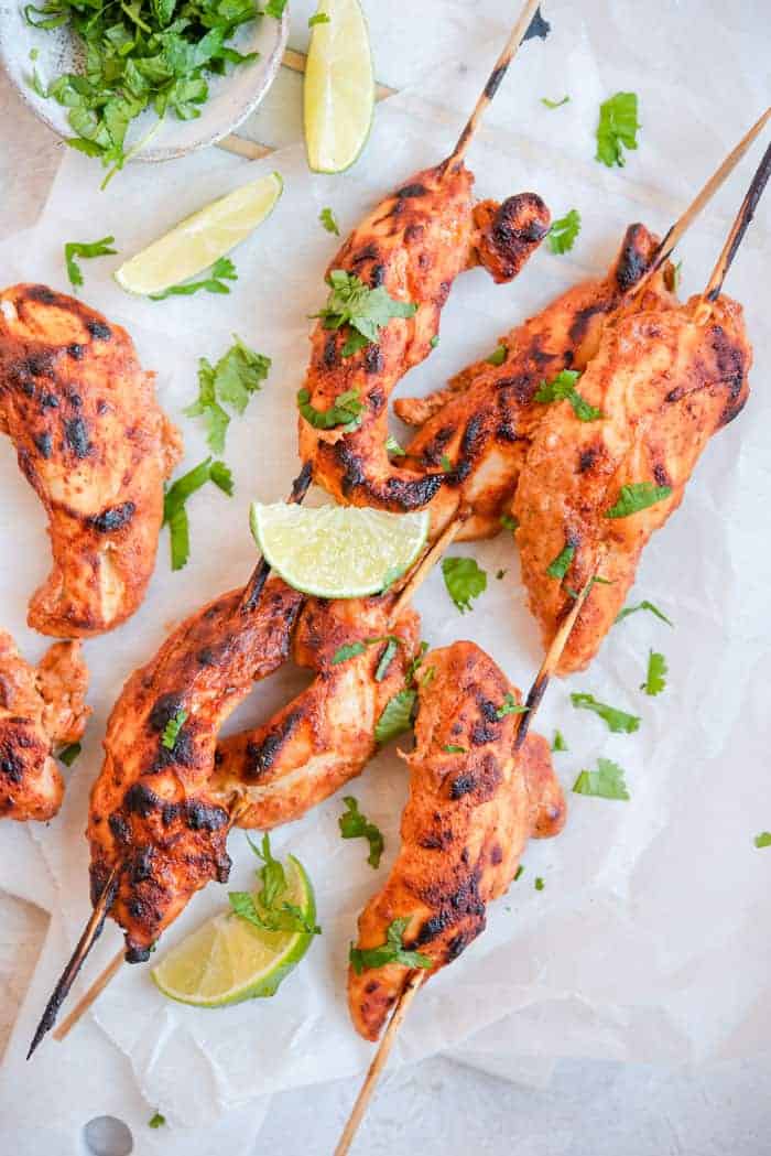An overhead shot of tandoori chicken skewers on a board with cilantro and lime wedges