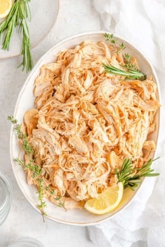 An overhead view of a large oval platter with Instant Pot shredded chicken topped with fresh rosemary and thyme and a lemon wedge.