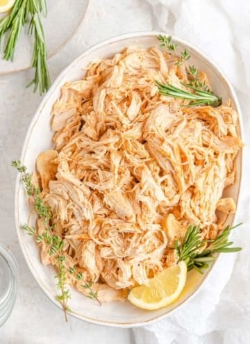 An overhead view of a large oval platter with Instant Pot shredded chicken topped with fresh rosemary and thyme and a lemon wedge.