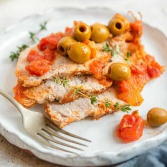 A close view of a plate with three pieces of sliced Instant Pot pork loin topped with olives, thyme, and tomatoes with a fork on the side.
