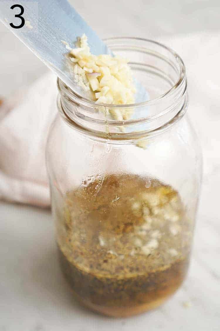 Minced garlic being added to a large jar of salad dressing.