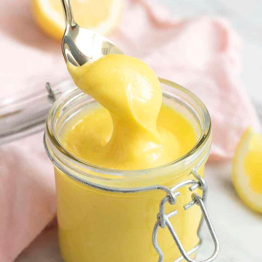 A thick homemade lemon curd getting spooned out of a glass jar.