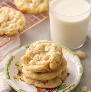 Three white chocolate macadamia nut cookies stacked on a plate alongside a glass of milk.
