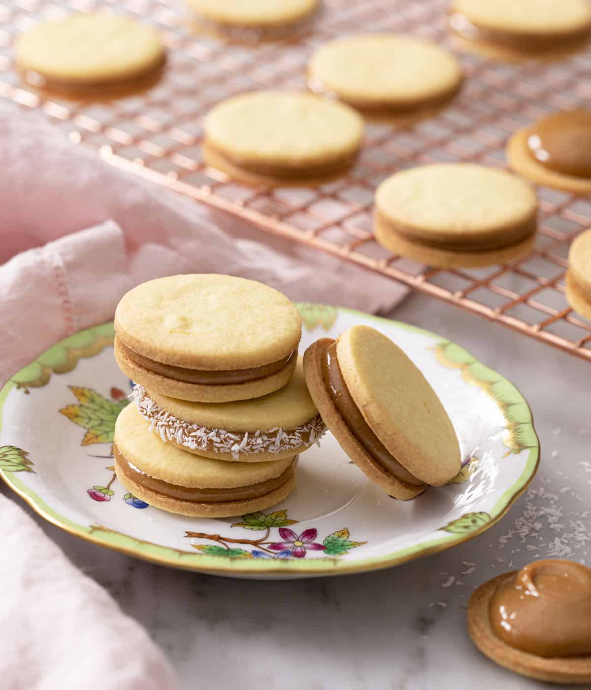 A group of alfajores cookies on a plate in front of a copper cooling rack filled with cookies. 