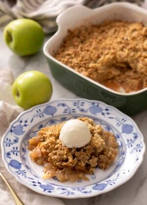 A portion of apple crisp with ice cream on a blue and white plate. A baking dish with more apple crisp in the background.