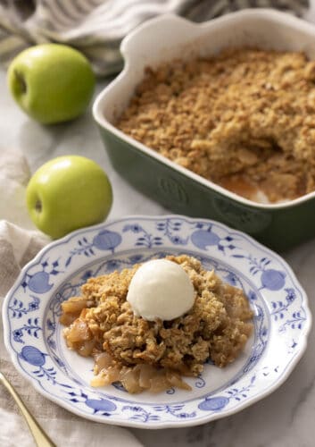 A portion of apple crisp with ice cream on a blue and white plate. A baking dish with more apple crisp in the background.