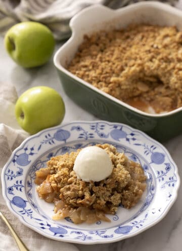A portion of apple crisp with ice cream on a blue and white plate. A baking dish with more apple crisp in the background.