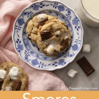 Two s'mores cookies on a blue and white plate nest to a glass of milk.
