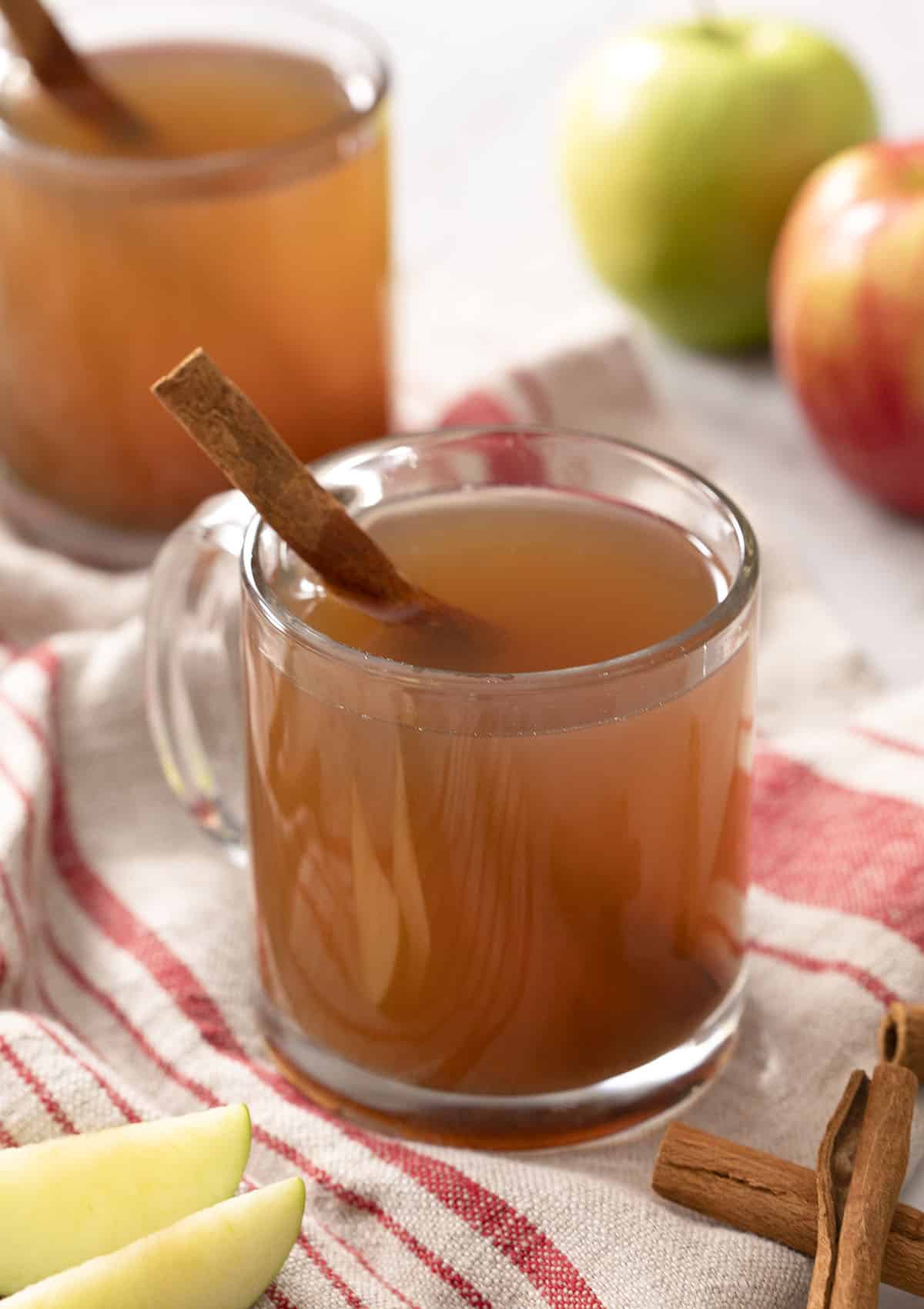 Two glasses of apple cider next to apples and cinnamon sticks.