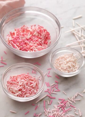 Pink homemade sprinkles in glass bowls on a marble counter.