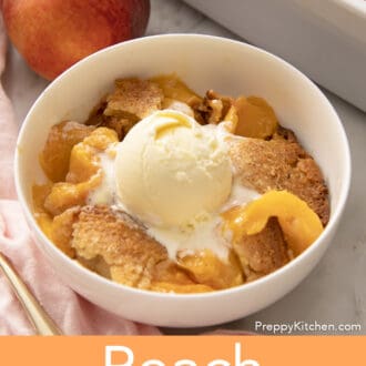 A white bowl of peach cobbler next to a peach.