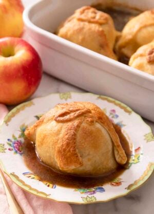 An apple dumpling on a porcelain plate. More apple dumplings in a baking dish in the background.