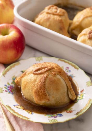 An apple dumpling on a porcelain plate. More apple dumplings in a baking dish in the background.