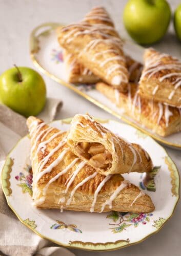 Two apple turnovers on a porcelain plate next to green apples. A bite taken out of a turnover on top.