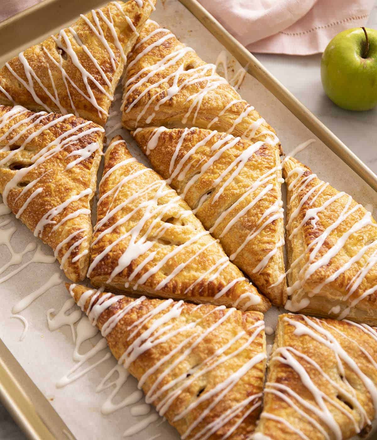 Eight apple turnovers arranged on a baking sheet with glaze drizzled on top.