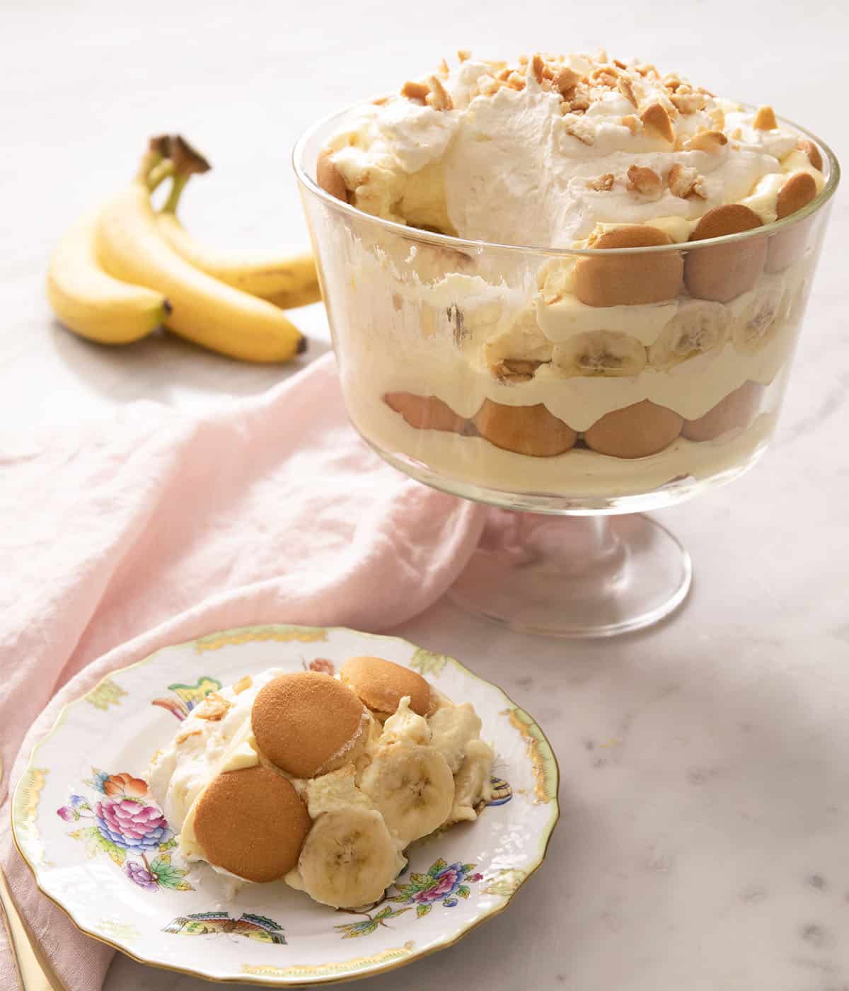 A serving of banana pudding on a porcelain plate next to a trifle dish.
