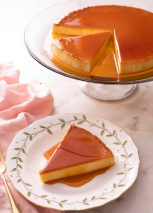 A slice of flan on a plate with the rest of the flan on a stand in the background.