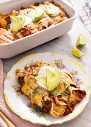 Chicken enchiladas on a plate with sour cream and avocado on top, with a casserole dish of enchiladas in the background.