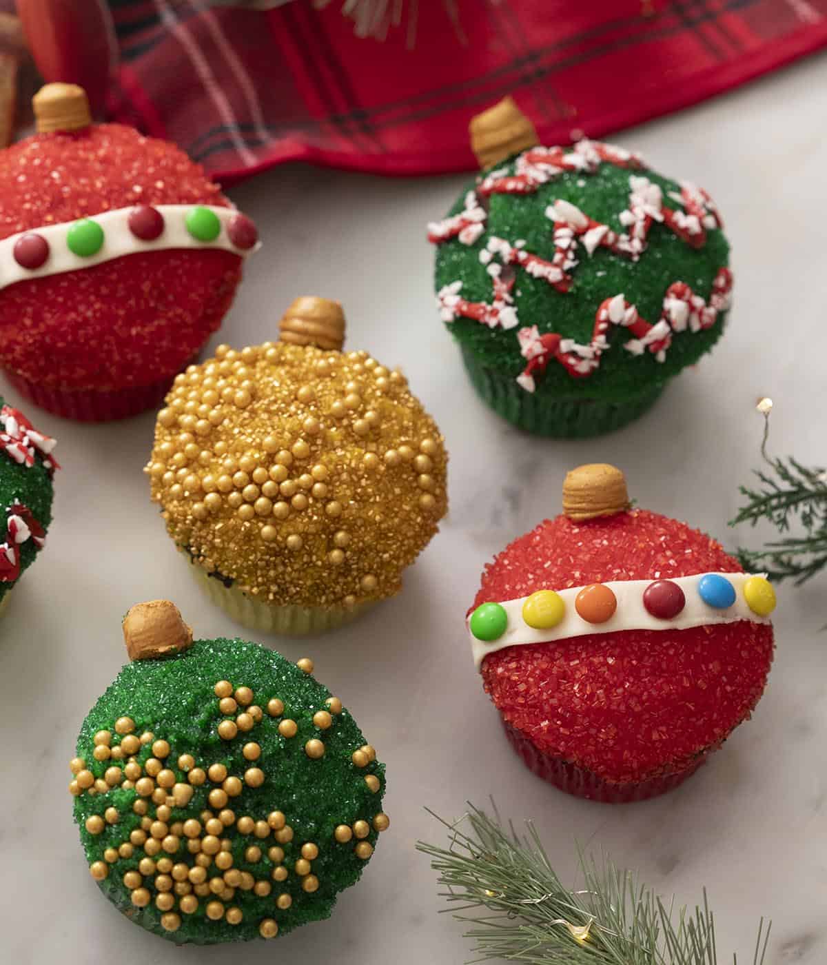 Christmas ornament cupcakes decorated with colorful sanding sugar and candies on a marble counter.