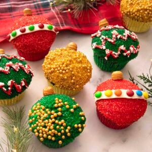 Christmas ornament cupcakes on a marble counter next to some pine sprigs.
