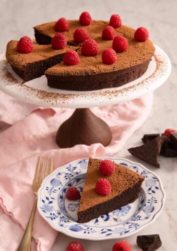 A flourless chocolate cake on a cake stand with a slice on a plate.