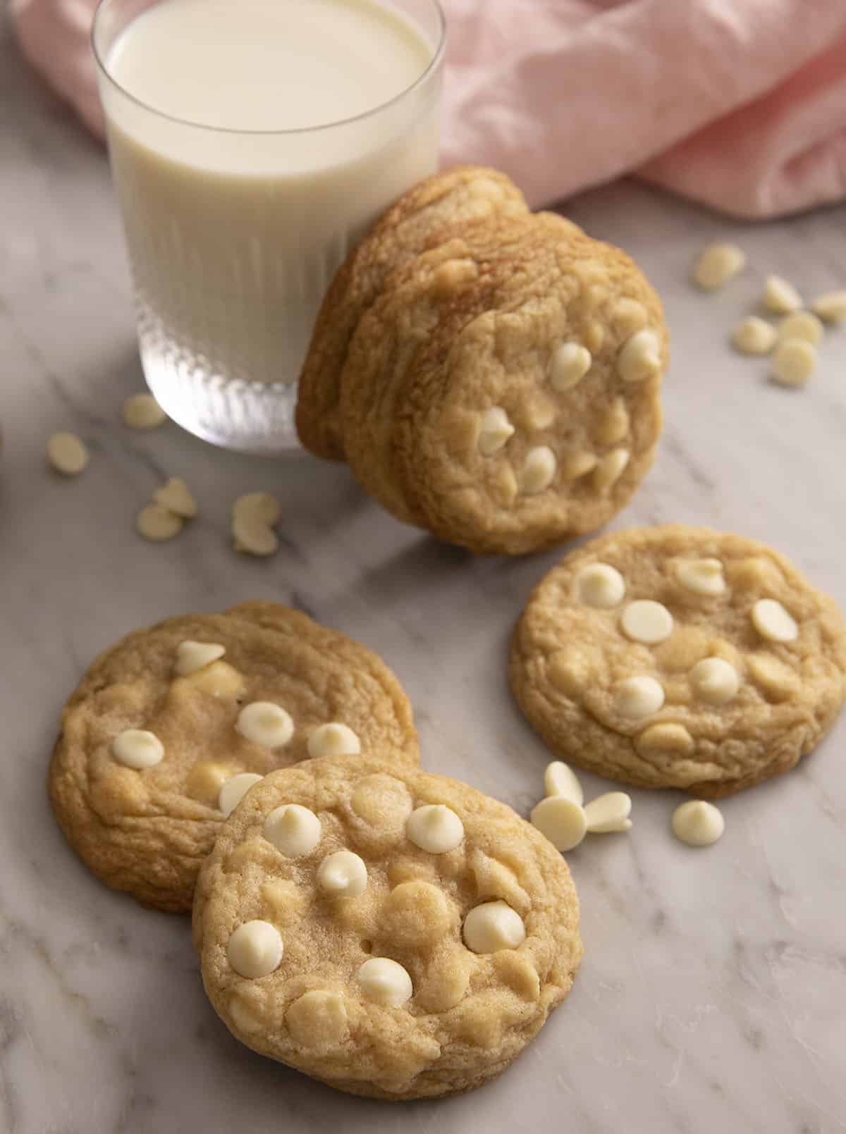 A close up of white chocolate chip cookies on a counter top with a glass of milk