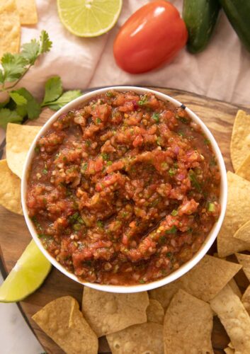 A close up of salsa in a bowl with tortilla chips and ingredients scattered around it.