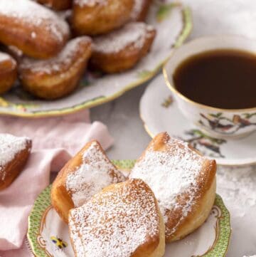 Three beignets on a plate with a cup of tea and platter of more beignets in the back.