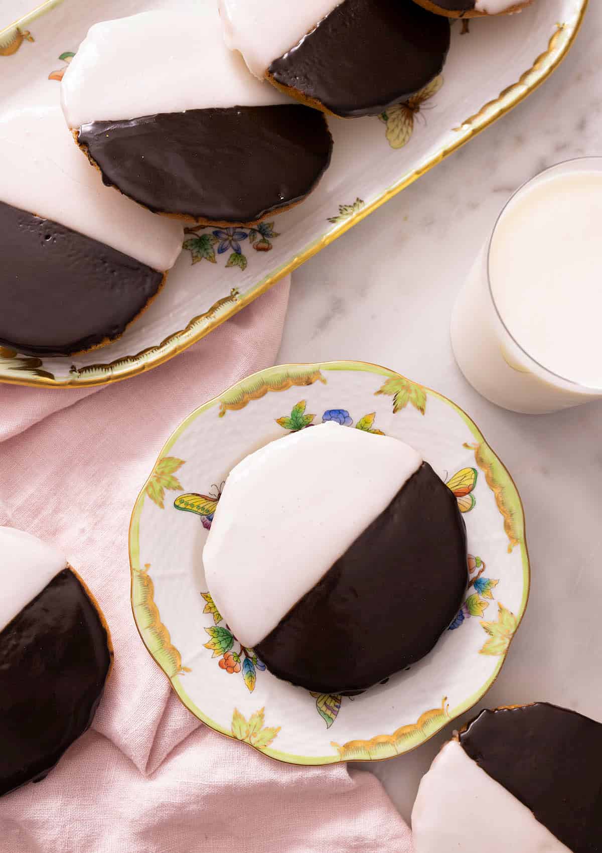 Overhead view of multiple black and white cookies with one in a plate beside a glass of milk.