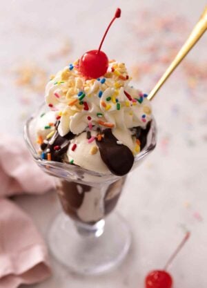 A glass of a hot fudge sundae made with vanilla ice cream with whipped cream, nuts, sprinkles, and a cherry on top.