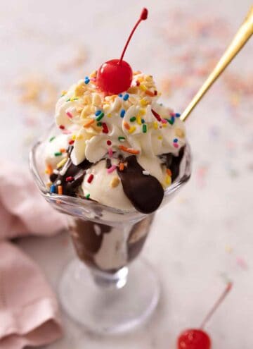 A glass of a hot fudge sundae made with vanilla ice cream with whipped cream, nuts, sprinkles, and a cherry on top.