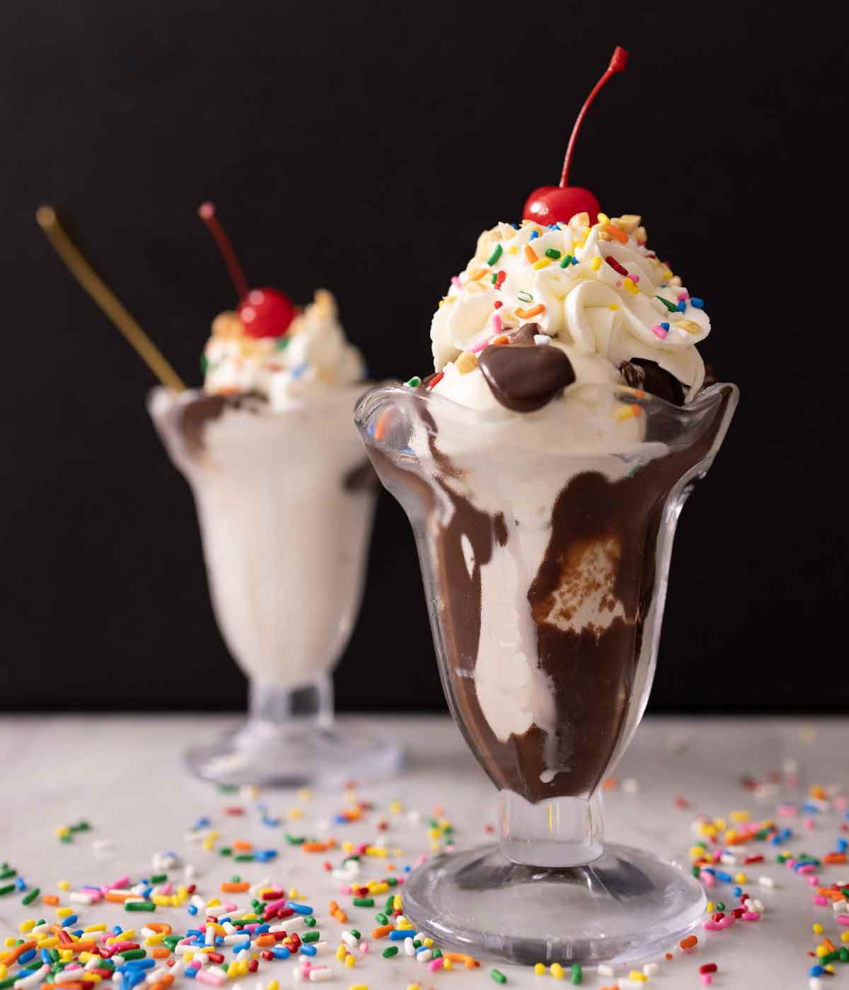 Two hot fudge sundae in a classic sundae glass with one in focus and with whipped cream on top and sprinkles on the table.
