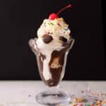 A classic sundae glass with a hot fudge sundae topped with sprinkles, whipped cream, and a cherry in front of a black background.