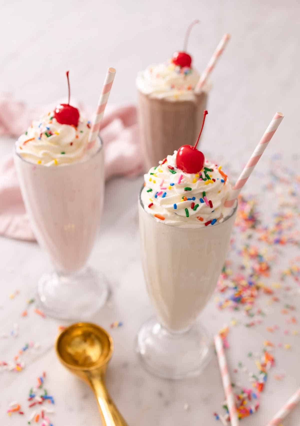 A vanilla milkshake, a strawberry milkshake, and a chocolate milkshake with toppings on top with sprinkles around the table.