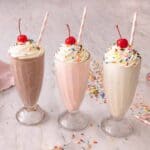Three glasses of milkshake in three different flavors, all topped with whipped cream, sprinkles, and a cherry.