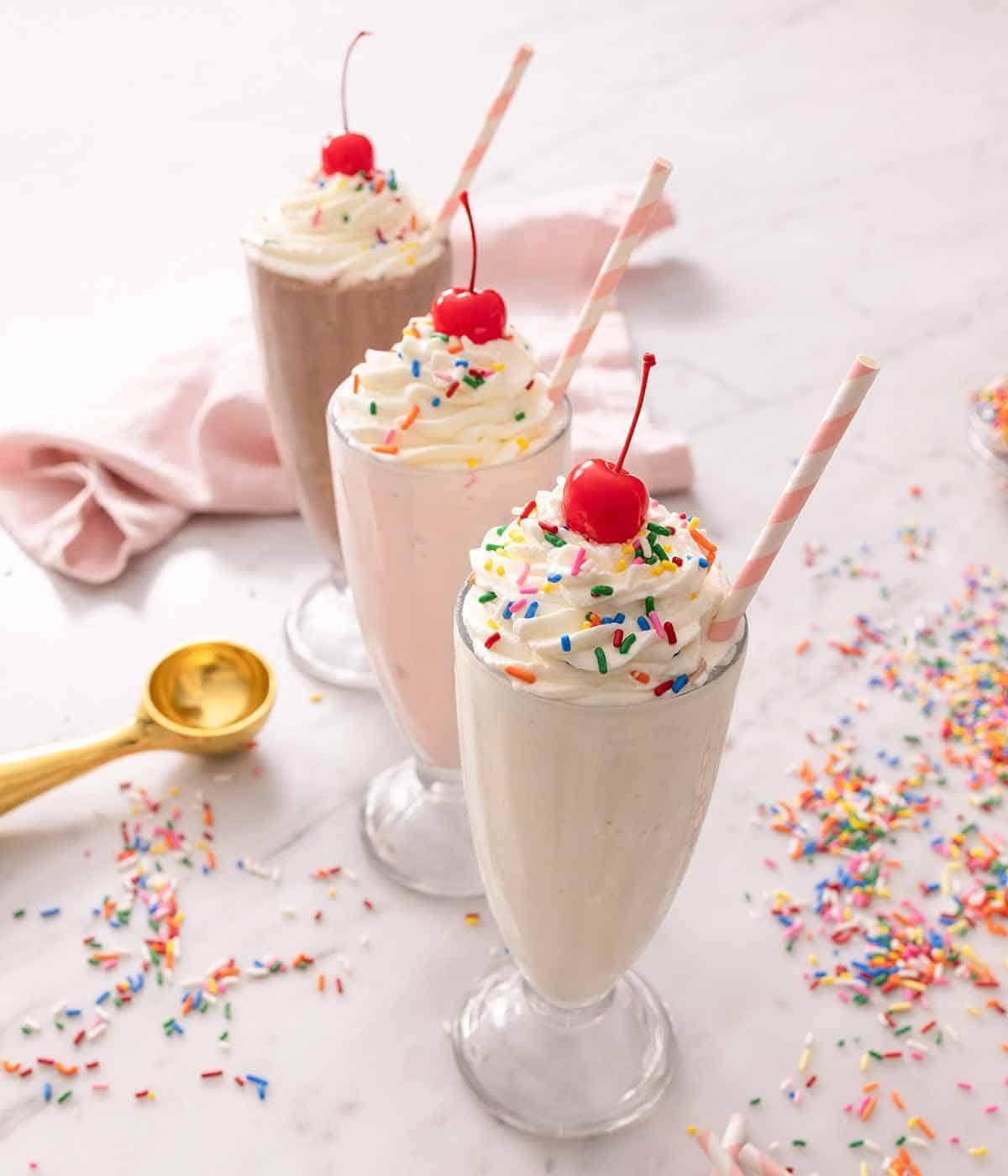 Three milkshakes: vanilla, strawberry, and chocolate flavored, all three with whipped cream, sprinkles, and a cherry on top.