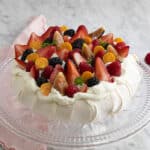 A Pavlova topped with whipped cream and berries on a glass cake stand