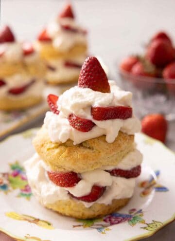 A plate and a long platter of strawberry shortcakes beside a bowl of strawberries with one plate in focus.