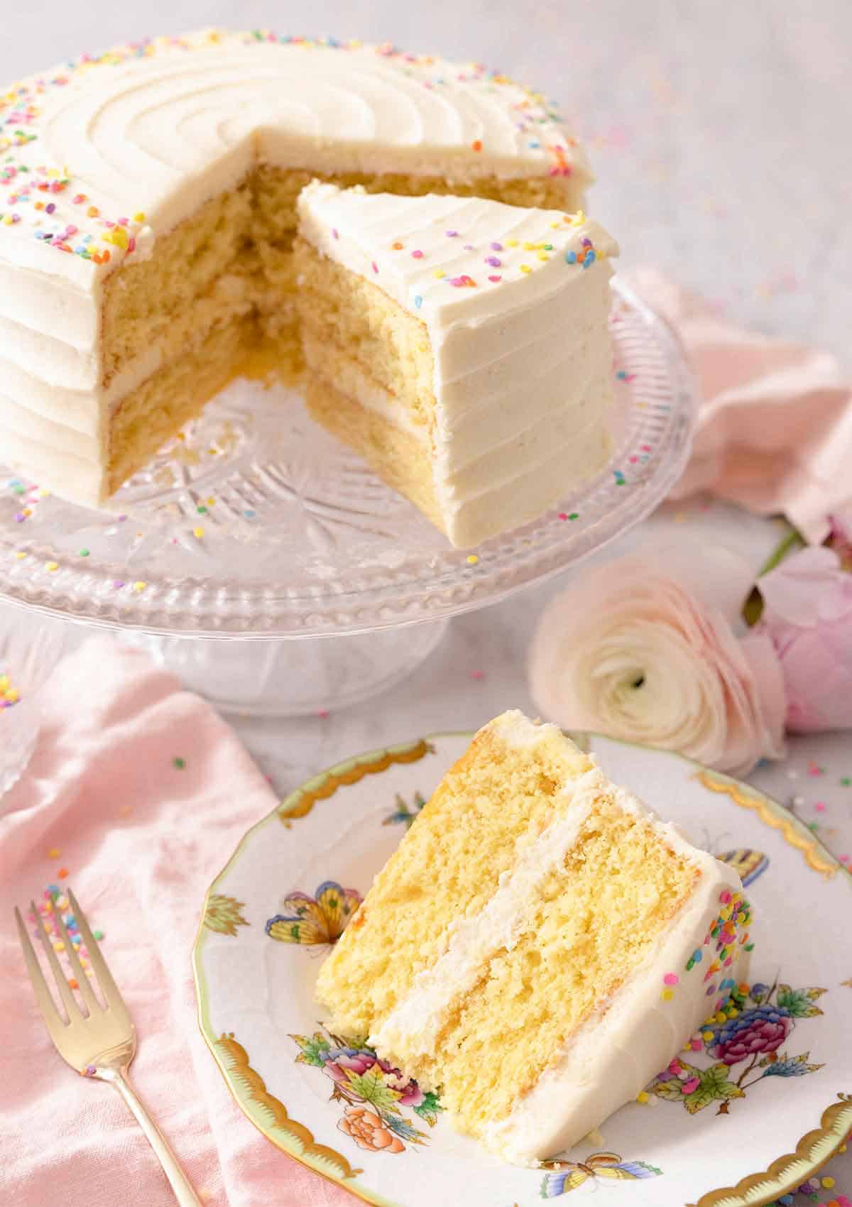 A slice of vanilla cake on a plate in front of the cake on a cake stand.