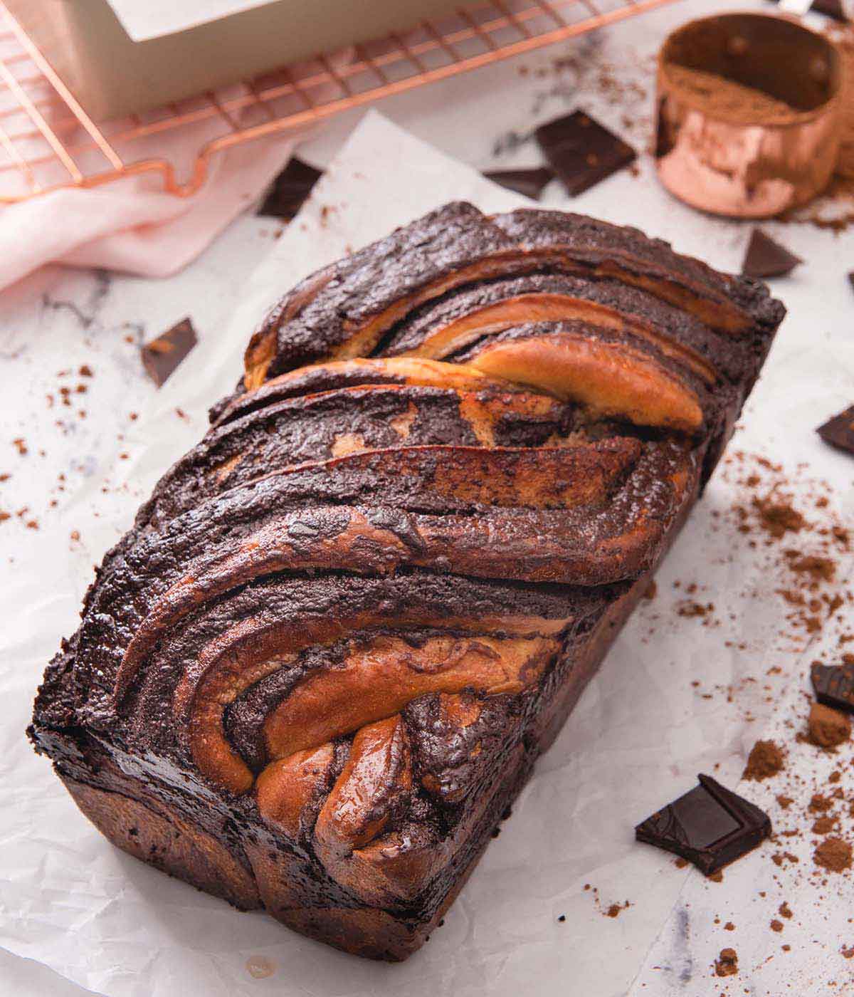 A loaf of chocolate babka on a sheet of parchment paper beside a rose gold measuring cup and wire rack.