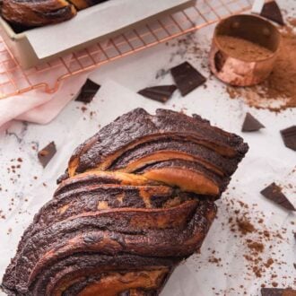 Pinterest graphic of babka with chopped chocolate and cocoa powder around it and a cooling rack with a second loaf in its pan, off to the side.