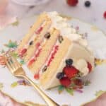 A slice of Chantilly cake lying on a plate on its side with berries on top and a fork beside it.