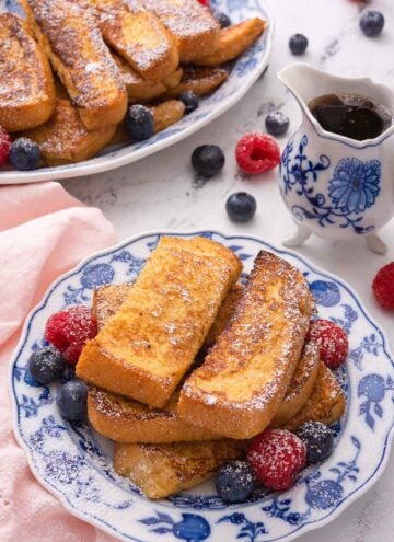 A plate of French toast sticks with powdered sugar on top and fresh berries around it. A serving platter with more is in the background along with syrup.