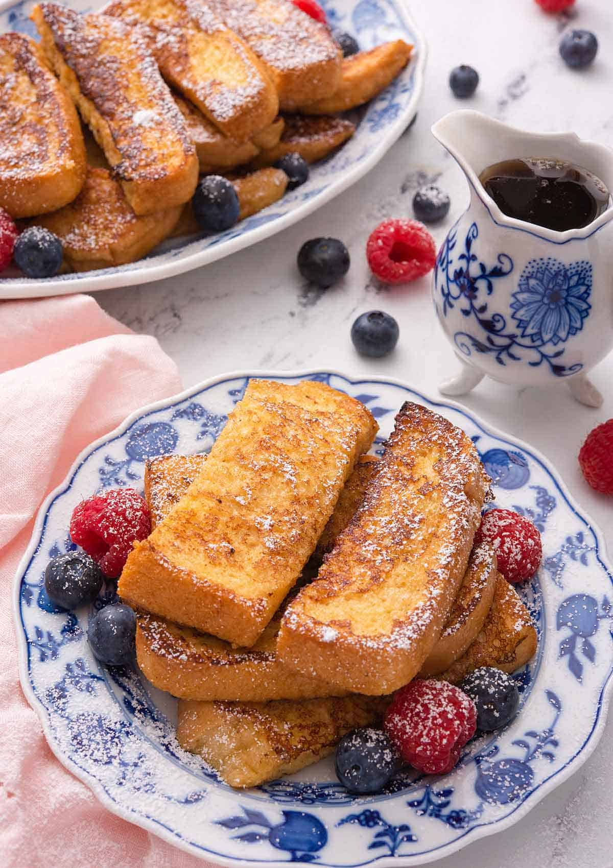 A plate of French toast sticks with powdered sugar on top and fresh berries around it. A serving platter with more is in the background along with syrup.