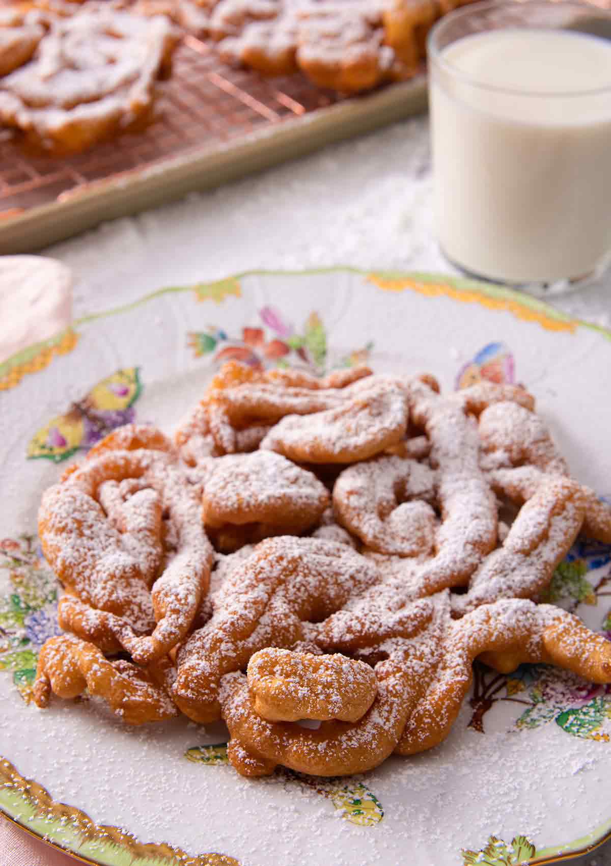 Funnel cake with a dusting of powdered sugar on a serving plate with a glass on milk in the background.